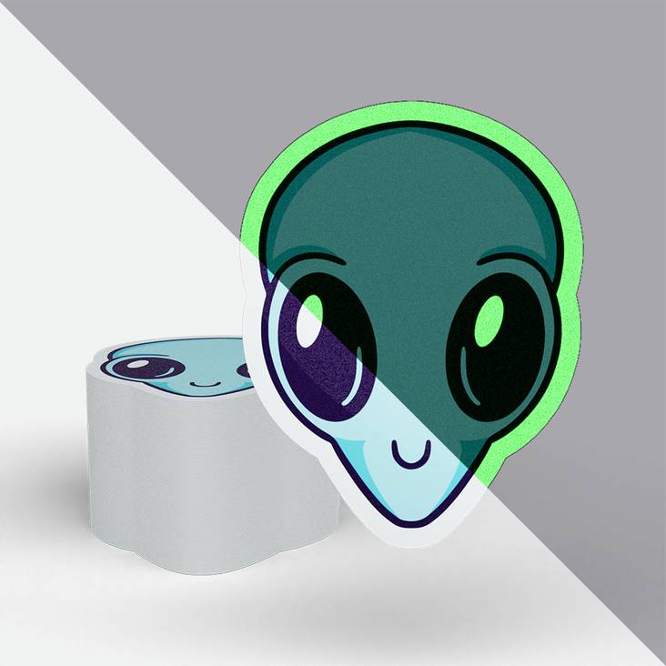 A stack of glow in the dark stickers printed by StickerGiant in the shape of an alien head - half of the picture is in dayligh and half of the picture is dark
