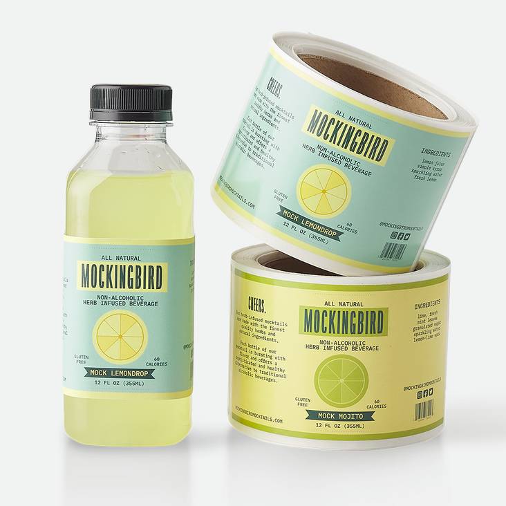 Two rolls of Mockingbird glossy paper labels printed by StickerGiant stacked next to a bottle of Lemondrop with glossy paper label applied to the bottle