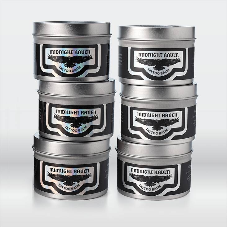 Stacks of metal tins with Midnight raven tattoo balm holographic labels printed by StickerGiant applied to the tins